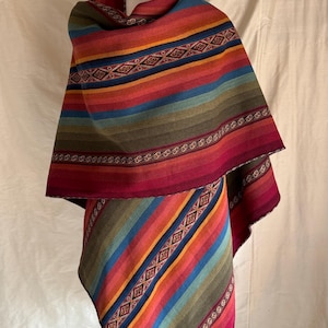 K&ouml;nnte beinhalten: Ein mehrfarbiger gestreifter Poncho auf einer Schaufensterpuppe. Der Poncho zeigt eine lebendige Farbpalette, darunter Rot, Orange, Gelb, Gr&uuml;n und Blau. Aufwendige geometrische Muster und dekorative Bord&uuml;ren verleihen dem Design Details. Das Kleidungsst&uuml;ck besteht aus einem weichen, gewebten Material.