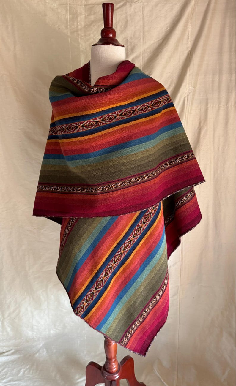 K&ouml;nnte beinhalten: Ein mehrfarbiger gestreifter Poncho auf einer Schaufensterpuppe. Der Poncho zeigt eine lebendige Farbpalette, darunter Rot, Orange, Gelb, Gr&uuml;n und Blau. Aufwendige geometrische Muster und dekorative Bord&uuml;ren verleihen dem Design Details. Das Kleidungsst&uuml;ck besteht aus einem weichen, gewebten Material.