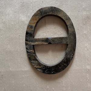 May include: Oval-shaped belt buckle with a dark, textured finish. The buckle has a horizontal bar across the center, and the material appears to have a natural, wood-like grain. The color is a mix of dark brown and tan.
