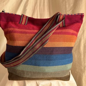 Könnte beinhalten: Eine farbenfrohe Tragetasche mit horizontalen Streifen in Rot-, Orange-, Lila-, Blau- und Grüntönen. Die Tasche hat einen passenden gestreiften Schulterriemen und einen Reißverschluss. Die Tasche ist aus einem gewebten Stoff gefertigt.