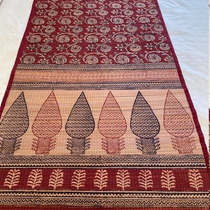 May include: A red and cream table runner with a floral pattern and a border of trees and leaves. The runner is made of a woven material.