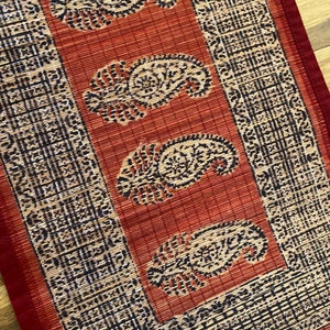 May include: A red and brown table runner with a paisley pattern. The runner has a black border with a geometric design.