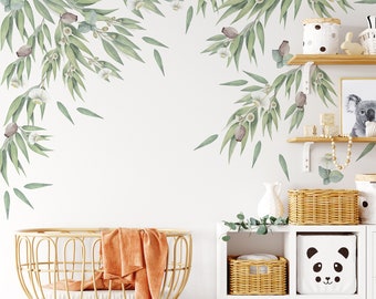 May include: A nursery room with a white wall decorated with green eucalyptus leaves and white flowers. A wicker bassinet with an orange blanket sits in the foreground. A white shelf unit with wicker baskets and a panda-themed storage bin is in the background.
