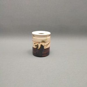 Può includere: Un piccolo vaso cilindrico in ceramica con una smaltatura bianca e disegni astratti marroni e neri. Il vaso ha un'apertura leggermente svasata nella parte superiore.