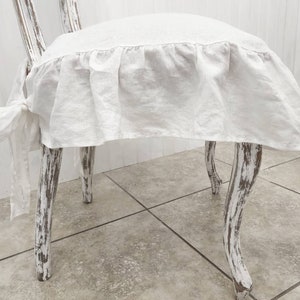 May include: A white linen chair cover featuring a ruffled skirt and side tie closures, fitted onto a distressed white wooden chair with curved legs. The fabric has a visible weave texture and drapes over the seat, secured by fabric ties.