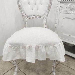 May include: A white shabby chic chair with a ruffled linen seat cushion. The chair features a tufted back and ornate, carved legs.  The chair is displayed against a white wood paneled wall. The chair's distressed white paint finish adds to its vintage charm.  The linen cushion provides a soft and comfortable seating surface. This elegant chair is perfect for adding a touch of rustic elegance to any room.