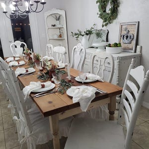 May include: A rectangular wooden dining table with a natural finish, surrounded by white distressed chairs with ruffled seat covers. The table is set with white plates, white cloth napkins, clear glassware, and small floral arrangements with greenery.