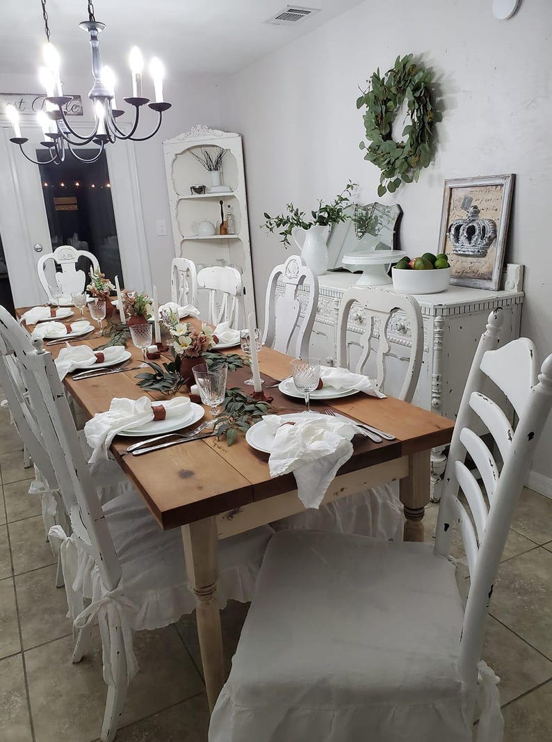 May include: A rectangular wooden dining table with a natural finish, surrounded by white distressed chairs with ruffled seat covers. The table is set with white plates, white cloth napkins, clear glassware, and small floral arrangements with greenery.