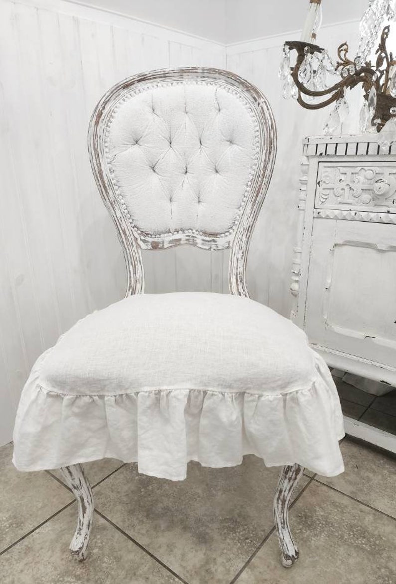 May include: A white shabby chic chair with a ruffled linen seat cushion. The chair features a tufted back and ornate, carved legs.  The chair is displayed against a white wood paneled wall. The chair's distressed white paint finish adds to its vintage charm.  The linen cushion provides a soft and comfortable seating surface. This elegant chair is perfect for adding a touch of rustic elegance to any room.