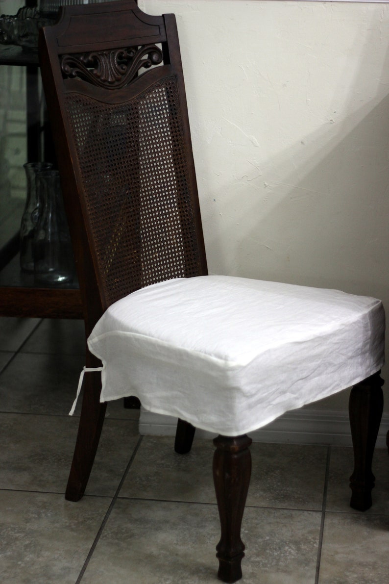Puede incluir: Una silla de madera de color marr&oacute;n oscuro con un respaldo alto que incluye un panel central de rejilla y detalles tallados en la parte superior. El asiento est&aacute; cubierto con una funda de tela blanca lisa sujeta con lazos laterales.