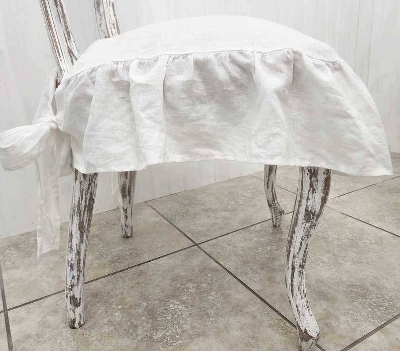 May include: A white linen chair cover featuring a ruffled skirt and side tie closures, fitted onto a distressed white wooden chair with curved legs. The fabric has a visible weave texture and drapes over the seat, secured by fabric ties.