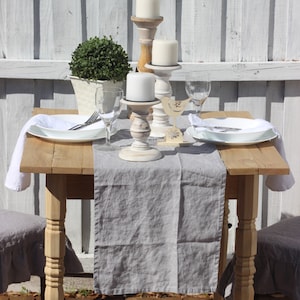 May include: A rustic wooden table with a gray linen tablecloth set for two. White candlesticks with white candles are on the table, along with two white plates and two wine glasses.