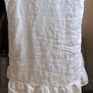 Puede incluir: Una funda de silla de lino blanco con una superficie de tela texturizada y un borde con volantes fruncidos en la parte inferior. La funda est&aacute; colocada sobre el respaldo de una silla de madera, mostrando un tejido natural y un aspecto suave.