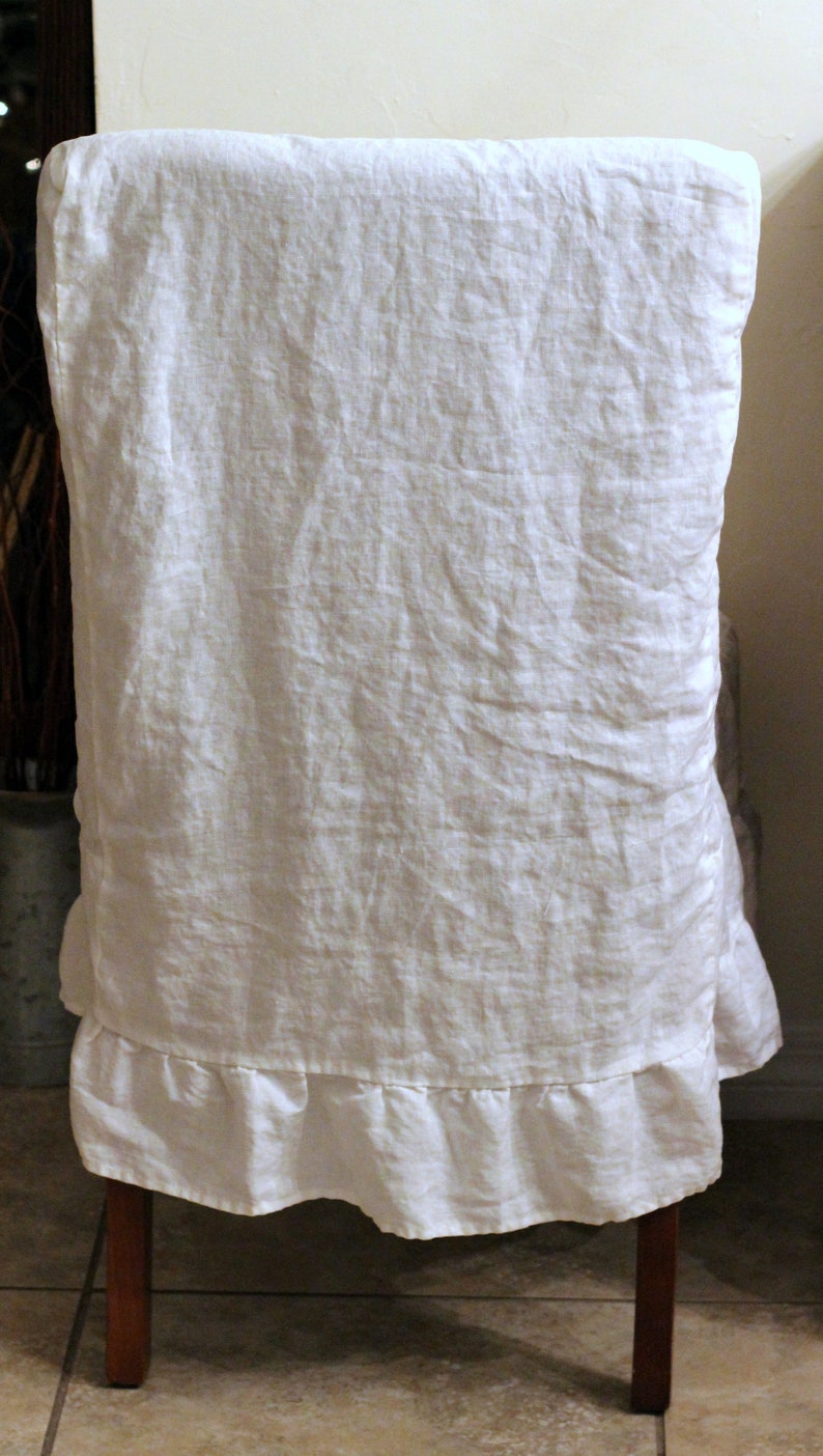 Puede incluir: Una funda de silla de lino blanco con una superficie de tela texturizada y un borde con volantes fruncidos en la parte inferior. La funda est&aacute; colocada sobre el respaldo de una silla de madera, mostrando un tejido natural y un aspecto suave.