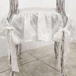 May include: A white linen chair cushion cover featuring a gathered ruffle skirt and long fabric ties on each side. The cover is secured to a wooden chair with a distressed white paint finish, showcasing a textured appearance and tied side bows.
