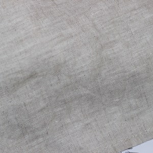 Puede incluir: Una vista de cerca de una tela de color beige claro natural con una textura tejida visible. El material presenta un patr&oacute;n de trama cruzada distintivo caracter&iacute;stico del lino, mostrando variaciones sutiles en el color del hilo y un tejido irregular.