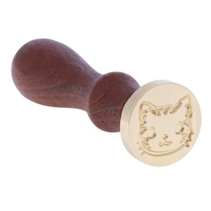 Può includere: Un timbro sigillo con manico in legno e testa del timbro rotonda in ottone. La testa del timbro presenta un disegno dettagliato di una faccia di gatto. Il manico è di colore marrone scuro.