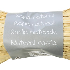 Può includere: Un rotolo di rafia naturale, di colore beige chiaro, ben avvolto. Un'etichetta grigia avvolta attorno al centro mostra il testo "Raffia natural" e "Natural raffia". Questa rafia è adatta per lavori artigianali e confezioni regalo.