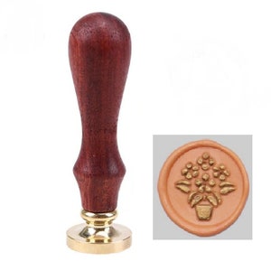 Peut inclure: Un tampon de cire avec un manche en bois brun foncé et une base en métal doré. L'empreinte du tampon présente un motif floral dans un pot. Le sceau est de couleur pêche clair.