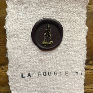 Könnte beinhalten: Handgeschöpftes Papier mit einem dunkelvioletten Wachssiegel, das ein goldenes Kerzenmotiv zeigt. Das Papier hat eine raue Oberfläche und die Worte "LA BOUGIE" sind in schwarzer Tinte gedruckt.
