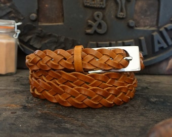 May include: A brown leather braided belt with a silver buckle. The belt is coiled and sitting on a wooden surface.