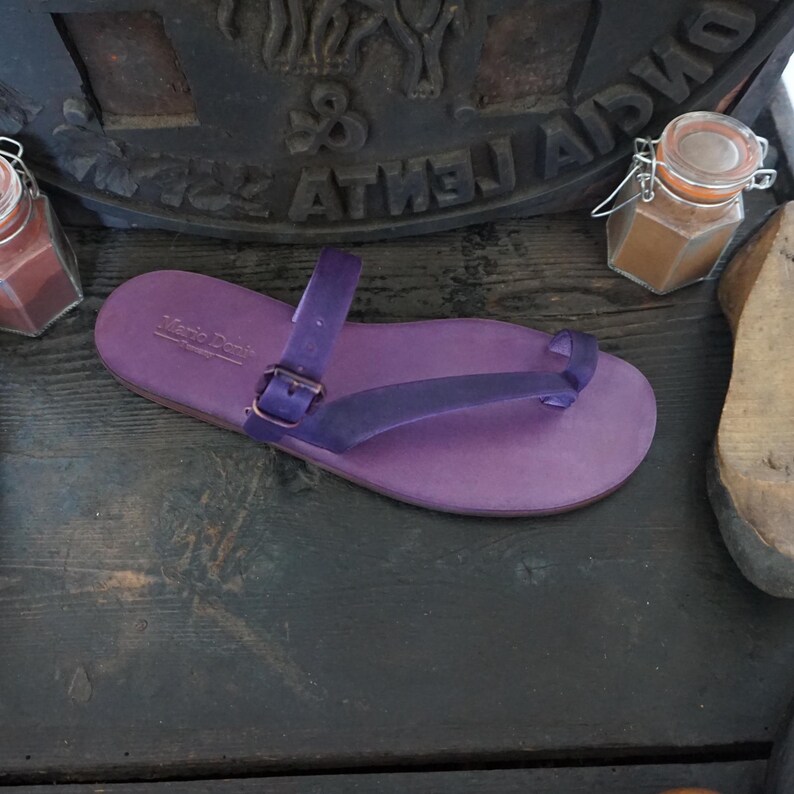 May include: Purple leather thong sandals with a buckle strap and a toe loop. The sandals are on a wooden surface.