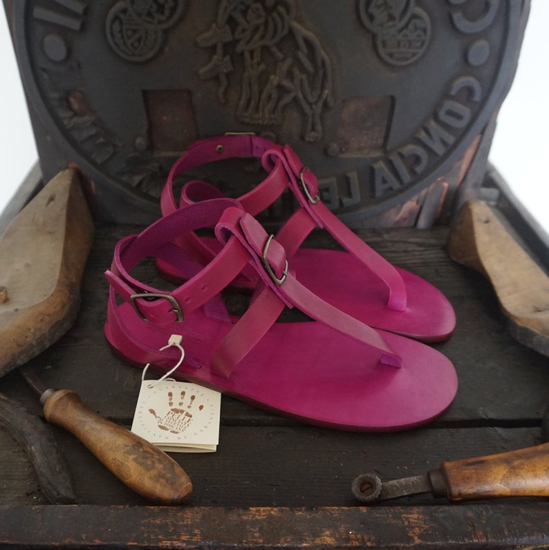 Pu&ograve; includere: Un paio di sandali in pelle rosa brillante con chiusura a fibbia e design a T. I sandali sono fatti a mano e hanno uno stile semplice ed elegante.