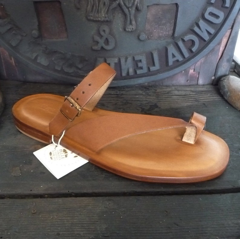 May include: Tan leather thong sandals with a buckle strap and a toe loop. The sandals have a thick, flat sole.