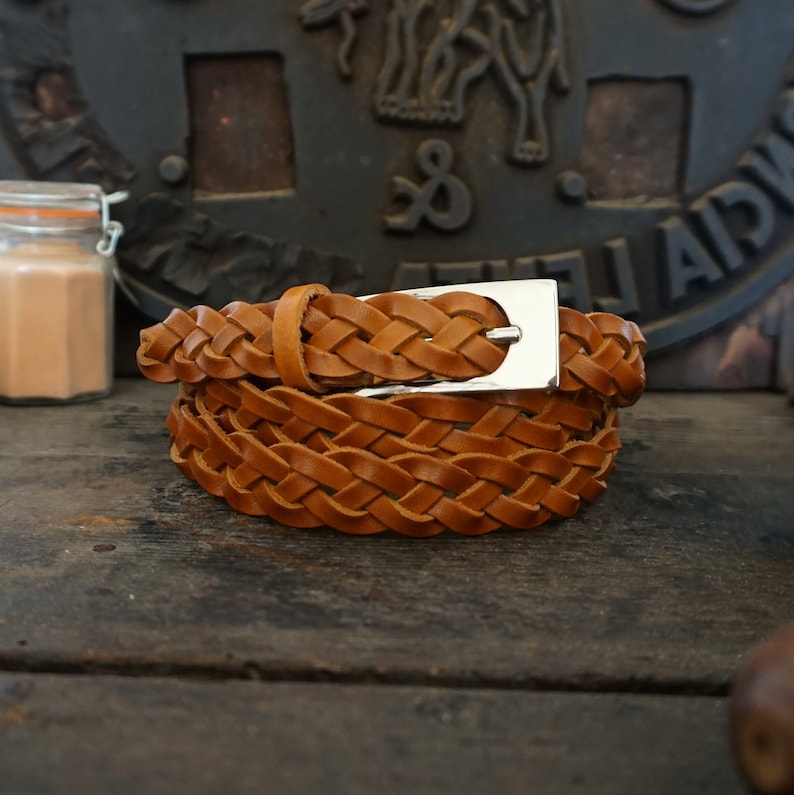 May include: A brown leather braided belt with a silver buckle. The belt is folded and lying on a wooden surface.