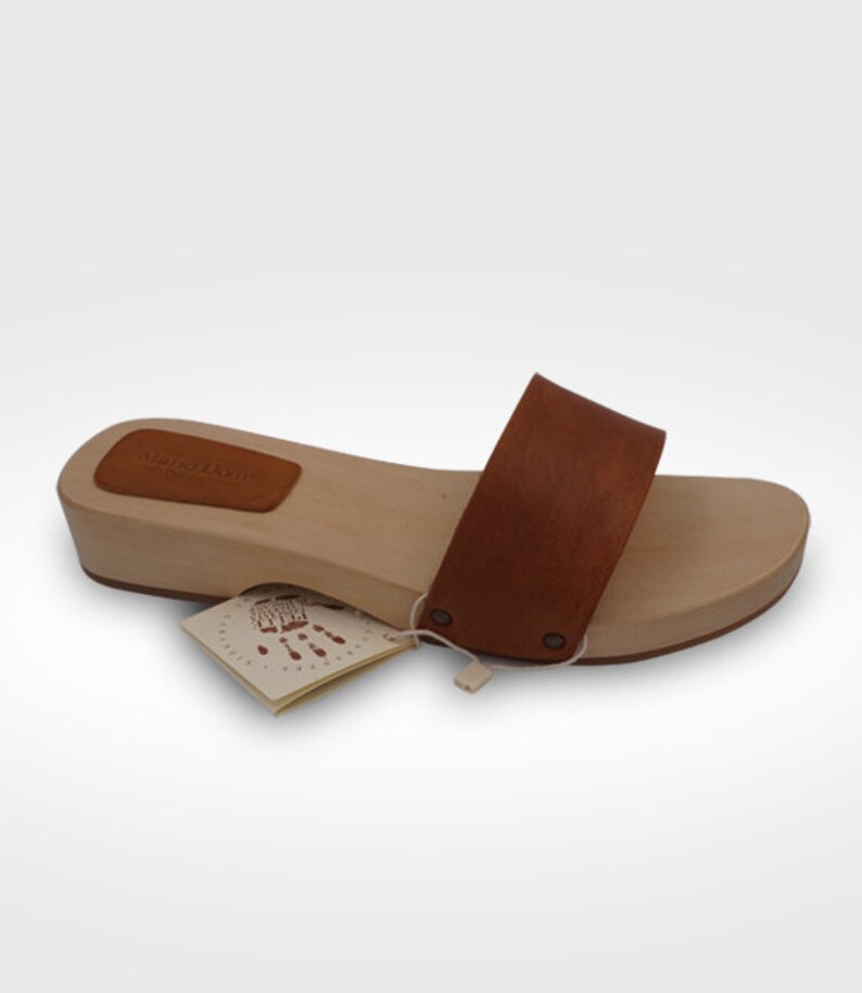 May include: A wooden clog with a brown leather strap and a light brown wooden base. The clog has a small brown leather patch on the heel with the text "Maria Dorn". A small tag with a handprint is attached.
