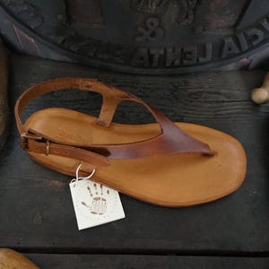 May include: Brown leather sandals with a buckle closure and a thong strap. The sandals have a hand-crafted look and are made with high-quality materials. The tag on the sandals reads "Pelle Conciata al Vegetale - Handmade in Tuscany".