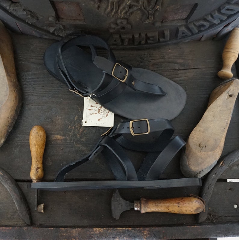 Pu&ograve; includere: Un paio di sandali in pelle nera con fibbie. I sandali sono su una superficie di legno con una variet&agrave; di strumenti.