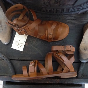 K&ouml;nnte beinhalten: Zwei Paar braune Ledersandalen mit mehreren Riemen und Schnallen. Die Sandalen haben eine flache Sohle und sind aus weichem, geschmeidigem Leder gefertigt.