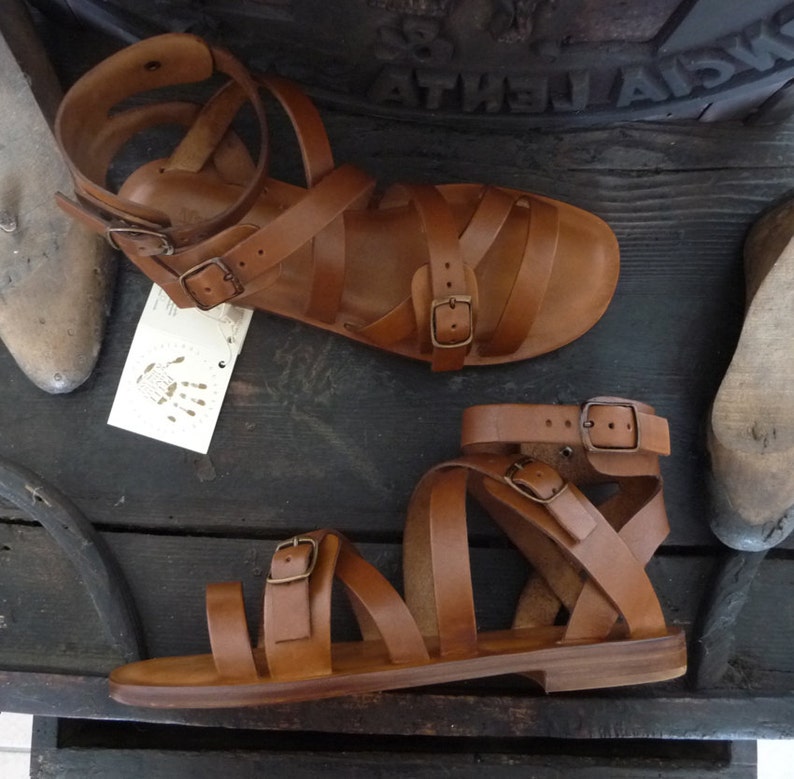 K&ouml;nnte beinhalten: Zwei Paar braune Ledersandalen mit mehreren Riemen und Schnallen. Die Sandalen haben eine flache Sohle und sind aus weichem, geschmeidigem Leder gefertigt.