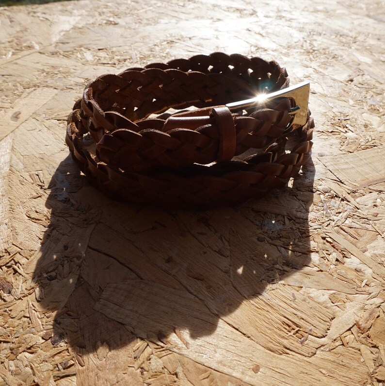 May include: A brown leather braided belt with a silver buckle. The belt is coiled up on a wooden surface.