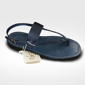 May include: A navy blue leather sandal featuring a thong-style front strap, a wide mid-foot band, and an adjustable ankle strap with a small metal buckle. A white paper tag with a handprint graphic and text is attached to the side of the sandal.