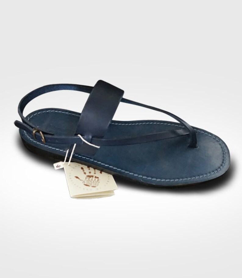 May include: A navy blue leather sandal featuring a thong-style front strap, a wide mid-foot band, and an adjustable ankle strap with a small metal buckle. A white paper tag with a handprint graphic and text is attached to the side of the sandal.