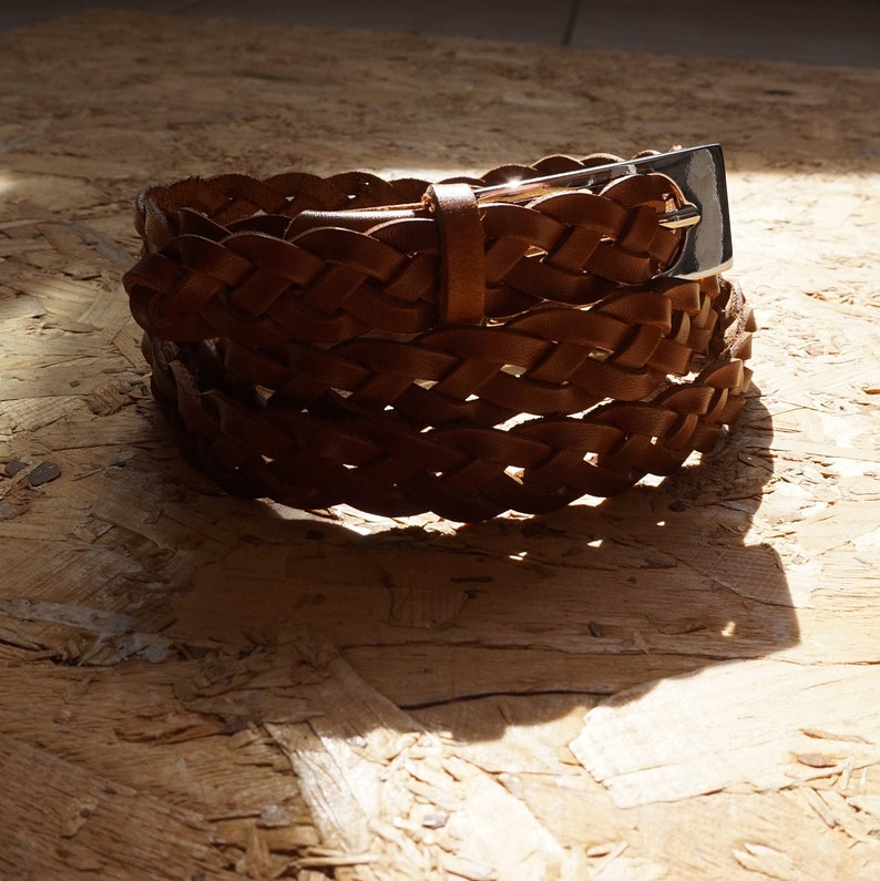 May include: A brown leather braided belt with a silver buckle. The belt is folded and lying on a wooden surface.