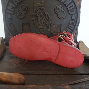 May include: Red leather sandals with a buckle closure and a textured sole. The sandals have a crescent moon logo on the sole.