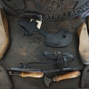 May include: Three pairs of black leather sandals with buckles and straps. The sandals are on a wooden surface with a metal object and a wooden tool.