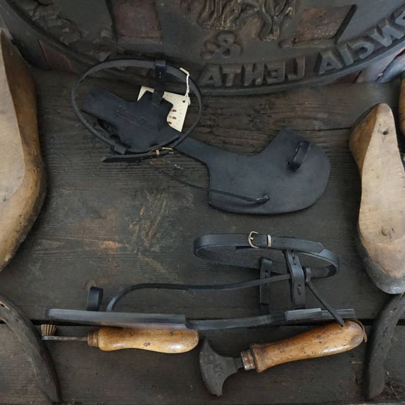 May include: Three pairs of black leather sandals with buckles and straps. The sandals are on a wooden surface with a metal object and a wooden tool.