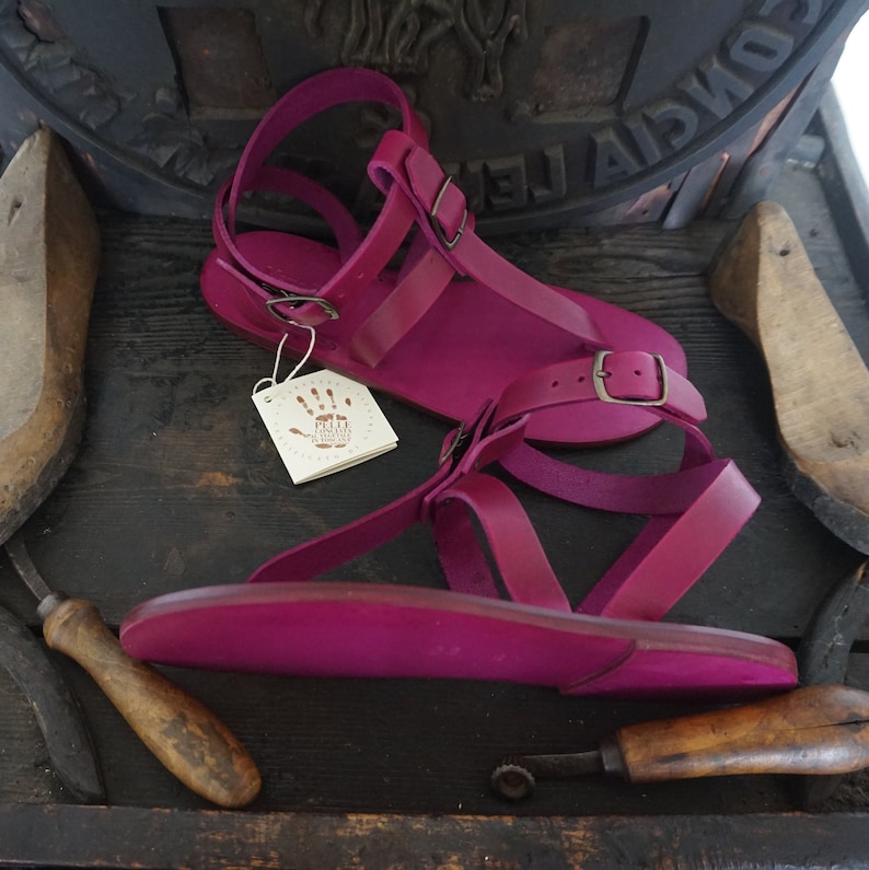 Pu&ograve; includere: Un paio di sandali in pelle rosa brillante con chiusura a fibbia. I sandali sono fatti a mano e hanno un design semplice e minimalista.