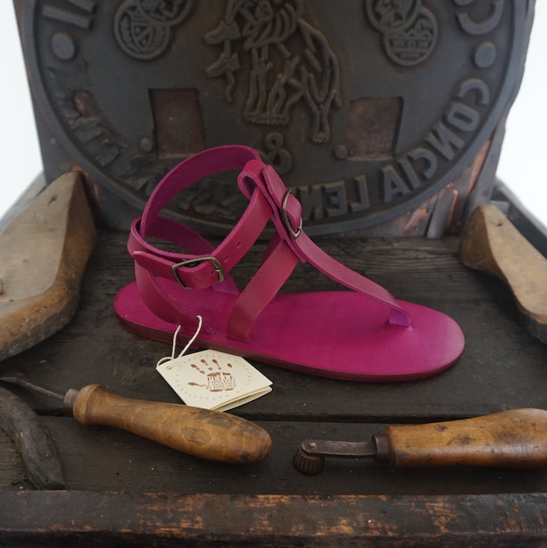Pu&ograve; includere: Un paio di sandali in pelle rosa brillante con cinturino con fibbia e cinturino per le dita. I sandali sono fatti a mano e hanno un design semplice ed elegante.
