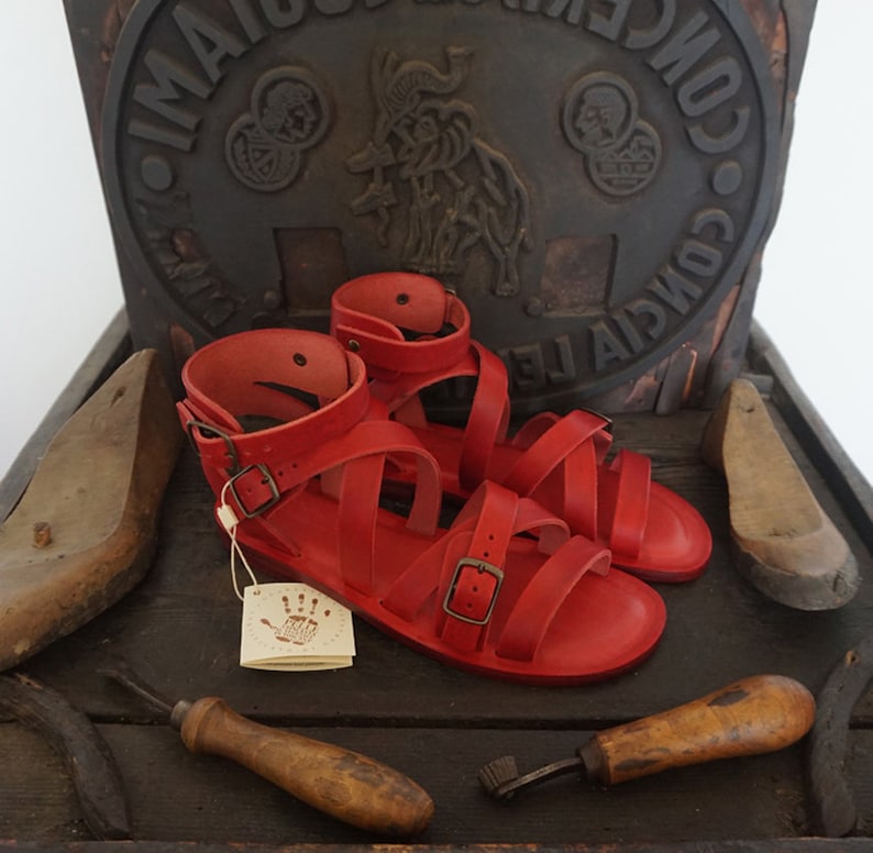 K&ouml;nnte beinhalten: Ein Paar rote Ledersandalen mit mehreren Riemen und Schnallen. Die Sandalen befinden sich auf einer Holzoberfl&auml;che mit anderen Lederwaren.