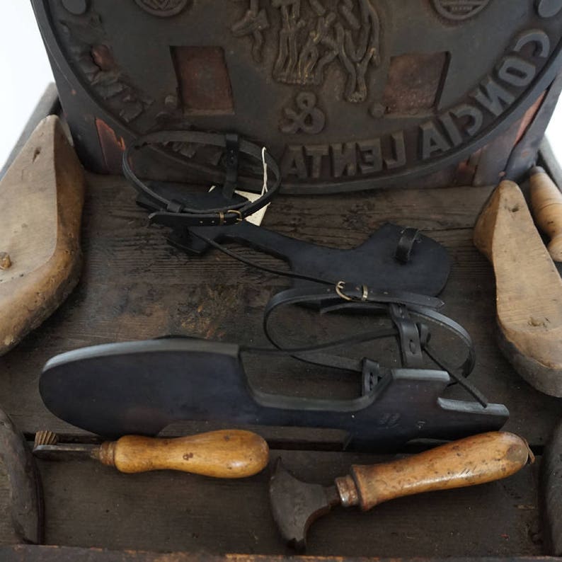 May include: Black leather sandals with straps and buckles, resting on a wooden surface with various tools, including a wooden mallet and a metal tool. The sandals are handmade and have a unique design.