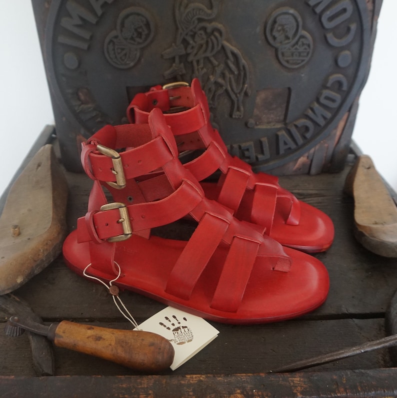 May include: A pair of red leather gladiator sandals with buckles. The sandals are on a wooden surface with a vintage stamp.