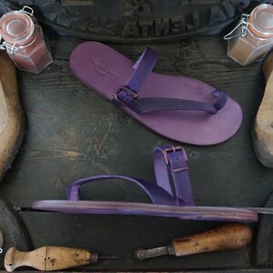 May include: Two pairs of purple leather sandals with buckles. The sandals have a thong style toe strap and a single strap that goes around the ankle.