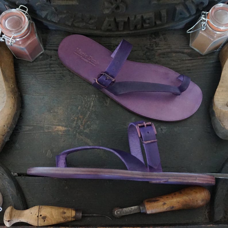 May include: Two pairs of purple leather sandals with buckles. The sandals have a thong style toe strap and a single strap that goes around the ankle.