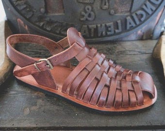 May include: A brown leather huarache sandal featuring a woven strap design across the upper, a closed toe, and an adjustable ankle strap with a metal buckle. The sandal has a flat sole and a smooth, polished finish on the leather straps.