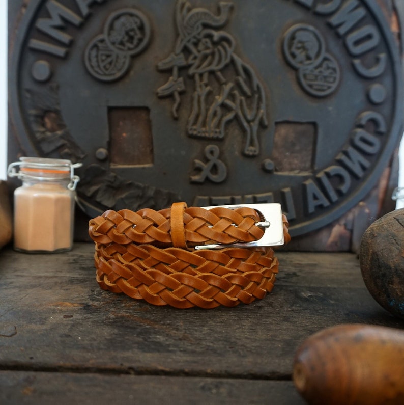 May include: A brown leather braided belt with a silver buckle. The belt is coiled and sitting on a wooden surface.